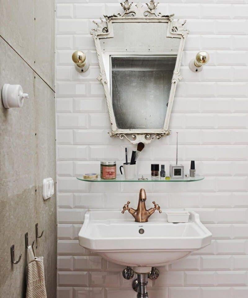 Koupelna kombinuje bílé obdélníkové obklady s jednoduchým umyvadlem a ozdobným zrcadlem. Baterie v teplém kovovém odstínu podtrhuje retro ladění. (Foto: A+Z Design Studio)