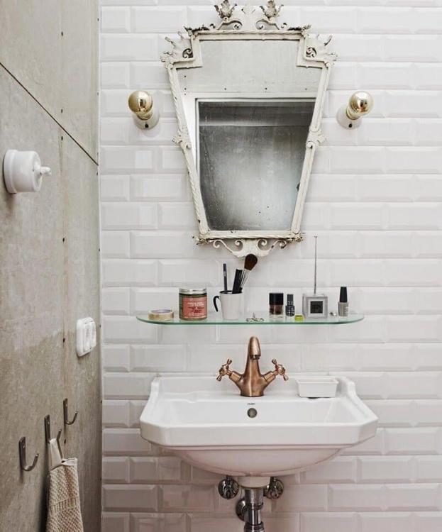 Koupelna kombinuje bílé obdélníkové obklady s jednoduchým umyvadlem a ozdobným zrcadlem. Baterie v teplém kovovém odstínu podtrhuje retro ladění. (Foto: A+Z Design Studio)