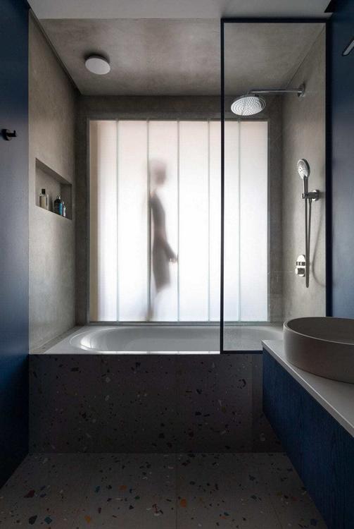 Matné profilované sklo propouští denní světlo k vaně a sprše. Šedé povrchy doplňuje podlaha s nepravidelnými barevnými úlomky. (Foto: Ivan Eroffev)